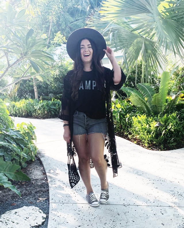 Girl in a hat with brown hair, waring a black shirt, jean shorts, and grey converse. Standing on a path with with greenery. 
