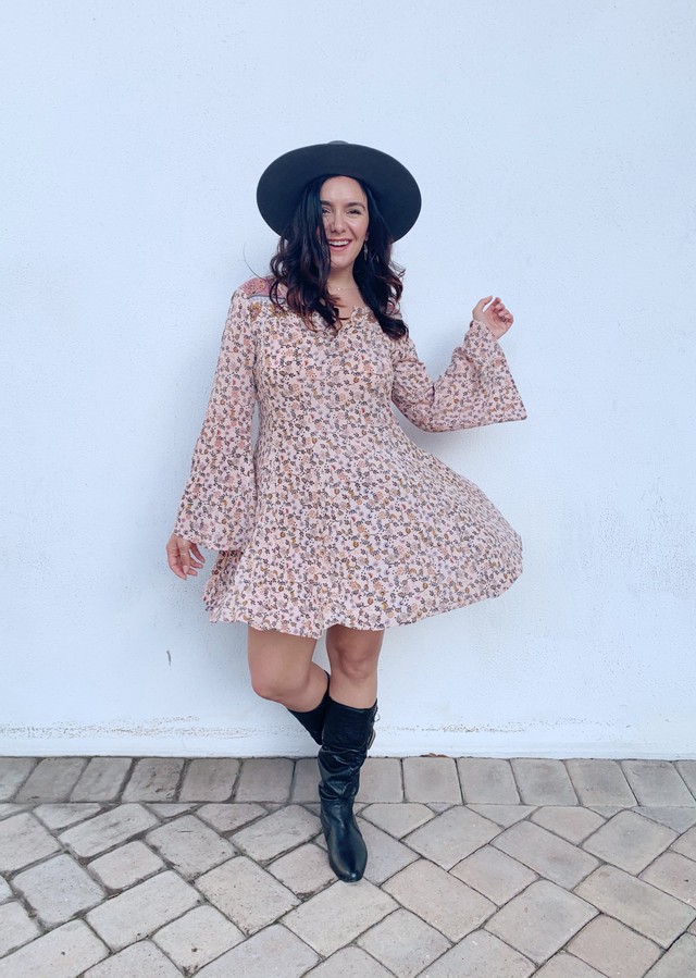 Girl with brown hair in a floral pink dress, black boots and hat smiling. 