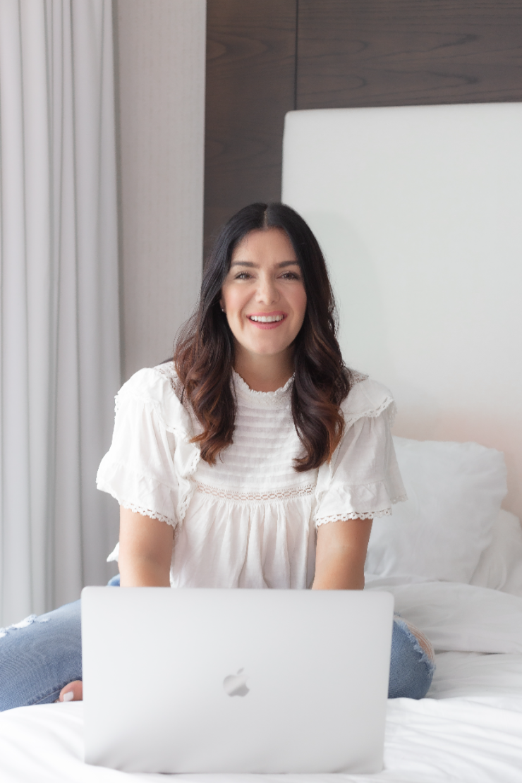 girl with brown hair, wearing a white shirt sitting on a bed on her laptop smiling. 