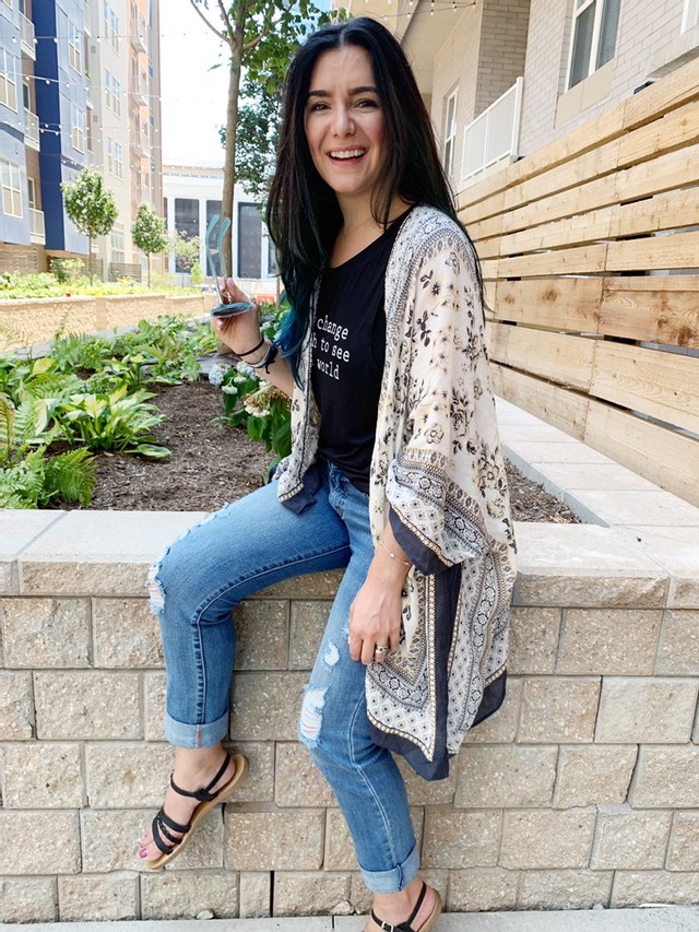Girl with brown hair, wearing jeans and black shirt and black sandals, sitting on a brick wall smiling.
